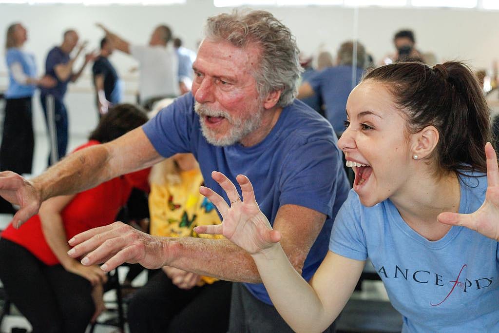 Two people wearing blue shirts, a young woman on the right next to an older man, make expressive faces with their arms raised in a Dance for PD program.