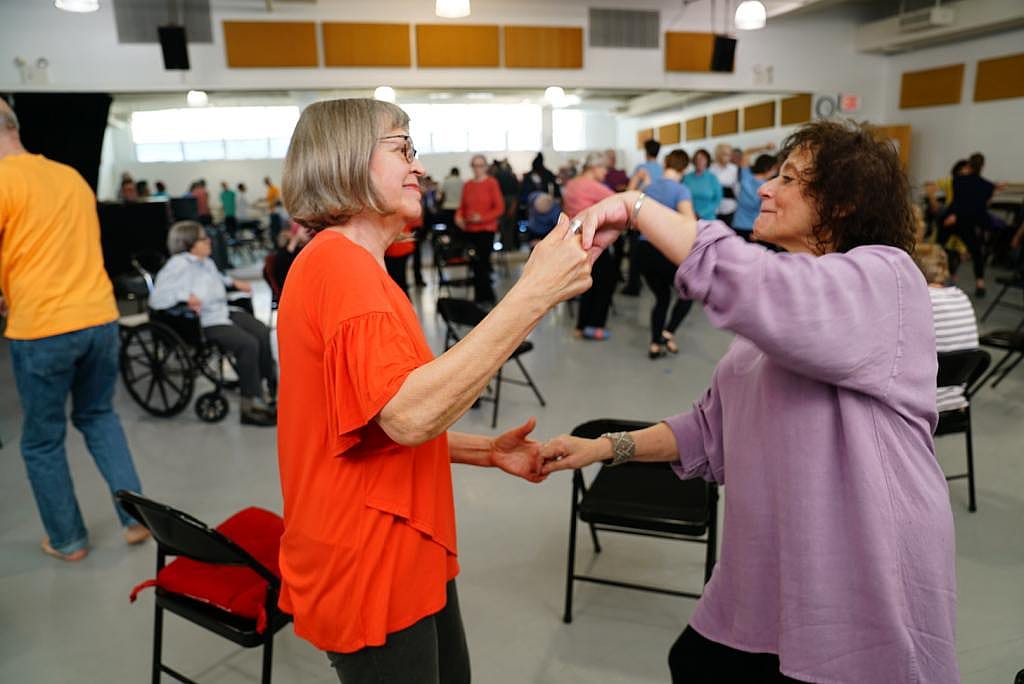 Side view of two people, one wearing orange and the other lavender, with joined hands swinging their arms as they dance, with many participants in the background also dancing.