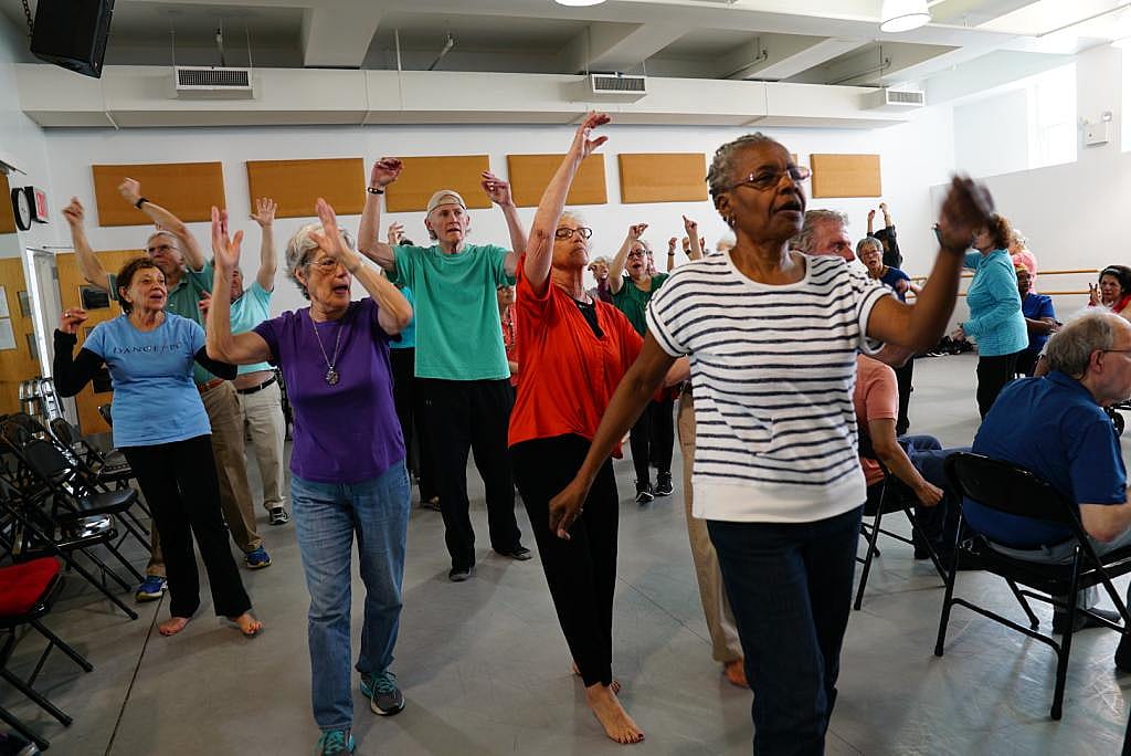 A woman in a black-and-white striped shirt stands, swinging her arms in front of a group of people in different colored shirts also dancing with their arms raised.
