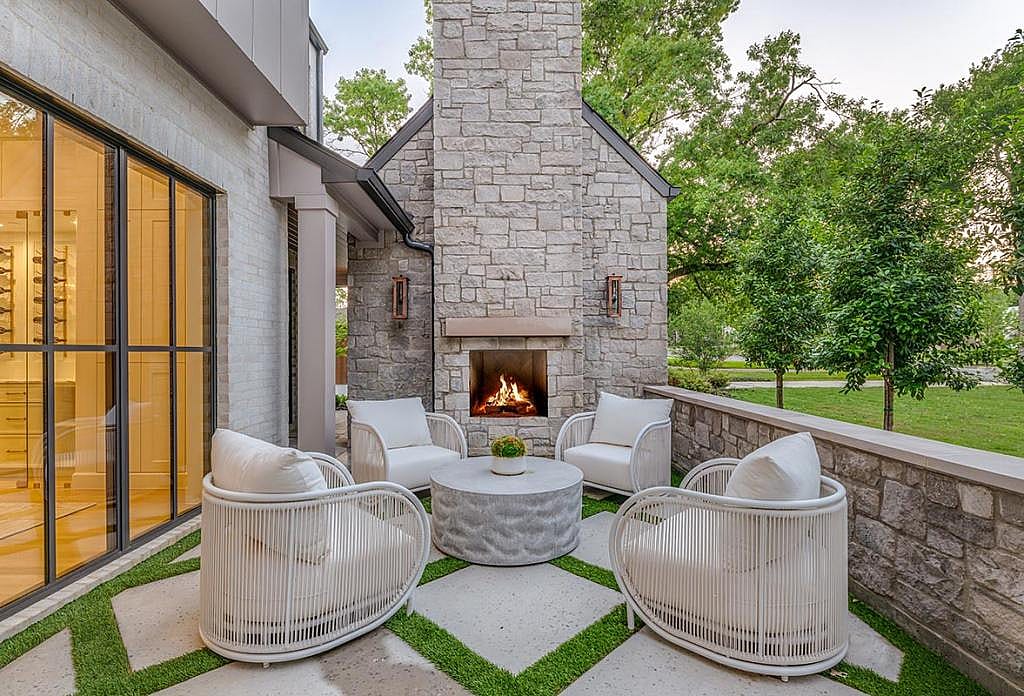 An outdoor patio layout and furnishings that include four large chairs centered around a table with a tall stone fireplace and chimney in the background. The patio includes large pavers with grass in between and has a stone retaining wall on one side to enclose it.