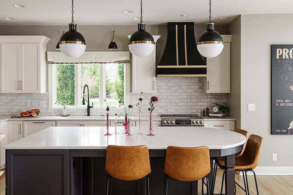 Kitchen with three round hanging light fixtures, a black and gold range hood, white tile and in the foreground a gently rounded kitchen counter stools around an island curved at one end.