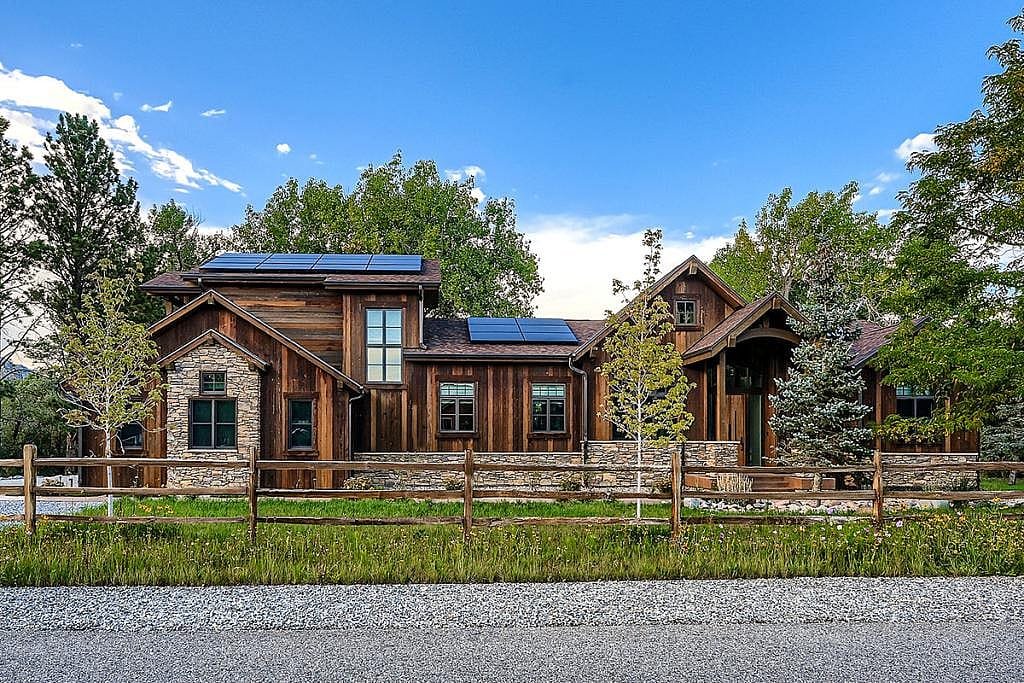 View of exterior of a house that is made with wooden and stone materials and has eco-friendly solar panels and native landscaping with a mixture of evergreen and deciduous trees planted in the yard.