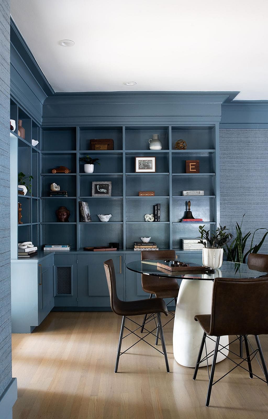 View of a room with blond wood floors and tall built-in open shelving painted in a light blue color featuring a cocoon-style room corner where a glass-topped table and four brown chairs offer a place to play board games or sit.