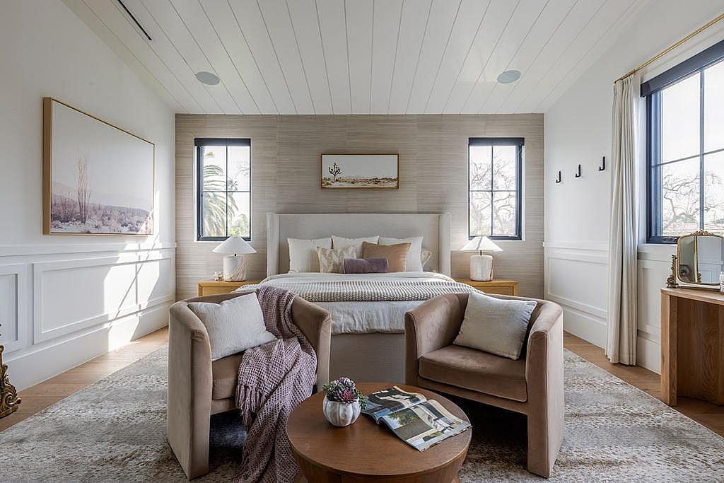 A bedroom decorated in warm neutral tones of pale blush, beige and mauve with two velvet chairs in the foreground in front of a bed, featuring an accent wall in a material with a luxurious feel.