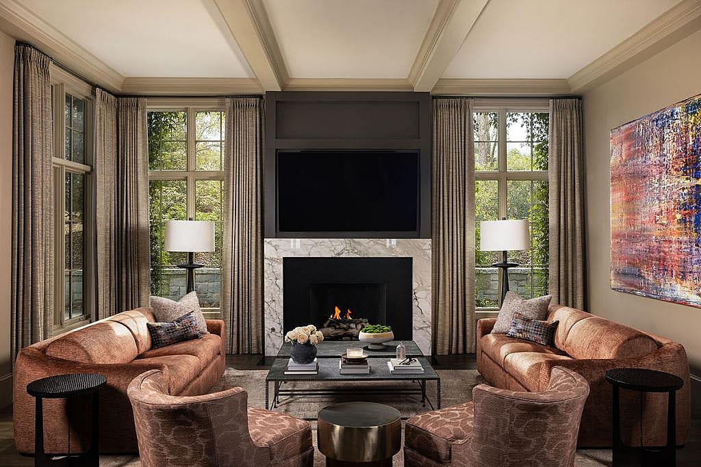 A living room with fireplace and tall windows featuring furniture in earthy rust hues with accent chairs, pillows, rugs and curtains.