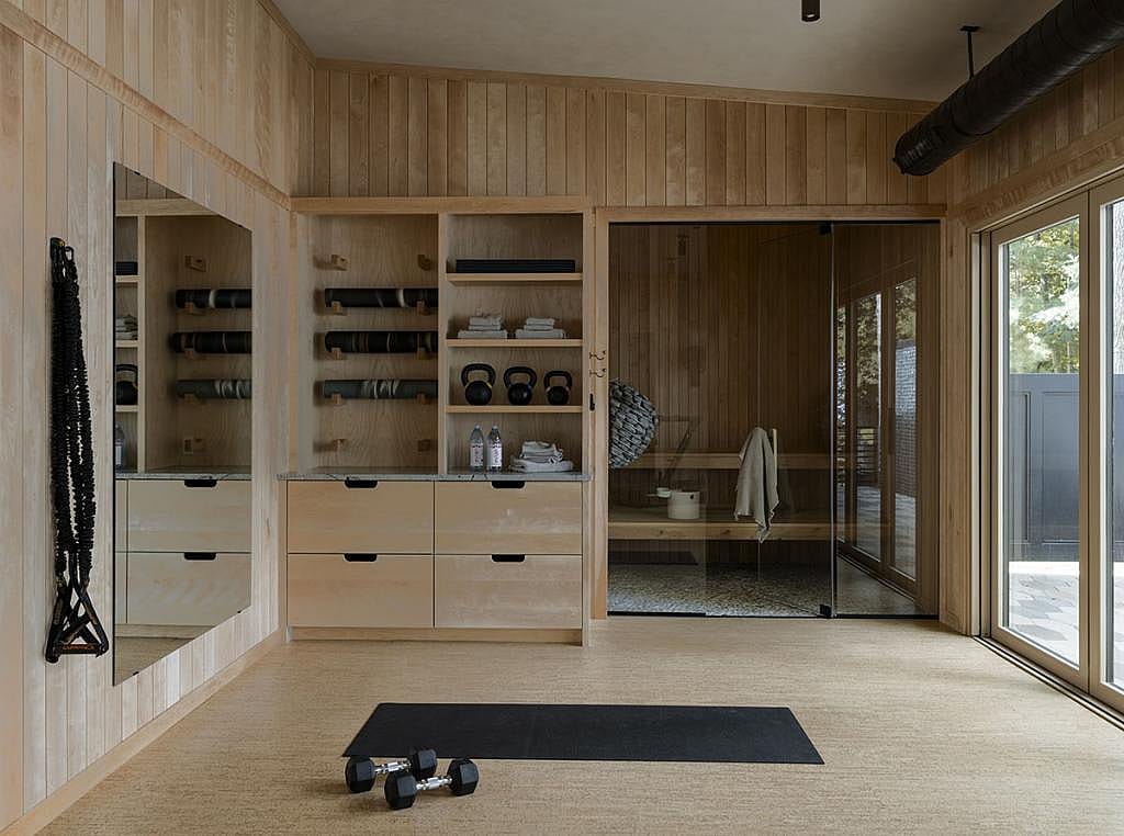 A wellness room with wood paneling on the walls, cabinet storage, a mirror for workouts, assorted weights and gym equipment displayed on shelving and a sauna in the back with glass doors.