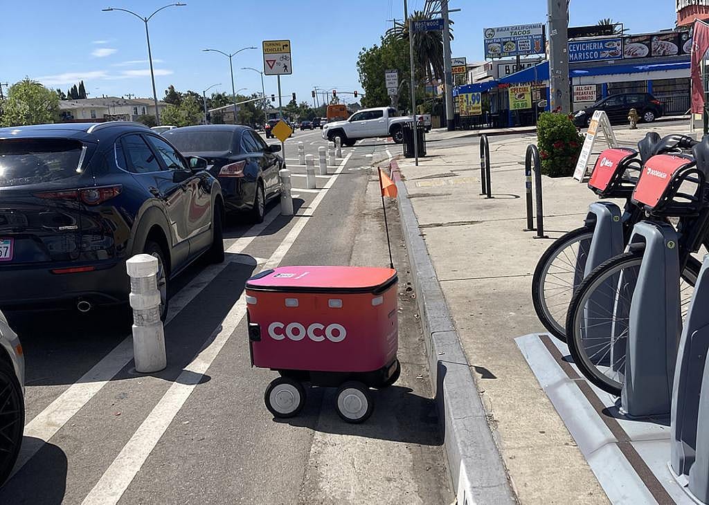 A pink 'coco' robot in the bike lane between parked cars and a bike rack.