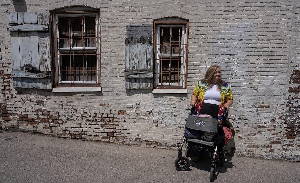 Cassandra Doyle holds the handles of a wheeled mobility standing in front of a whitewashed brick wall.