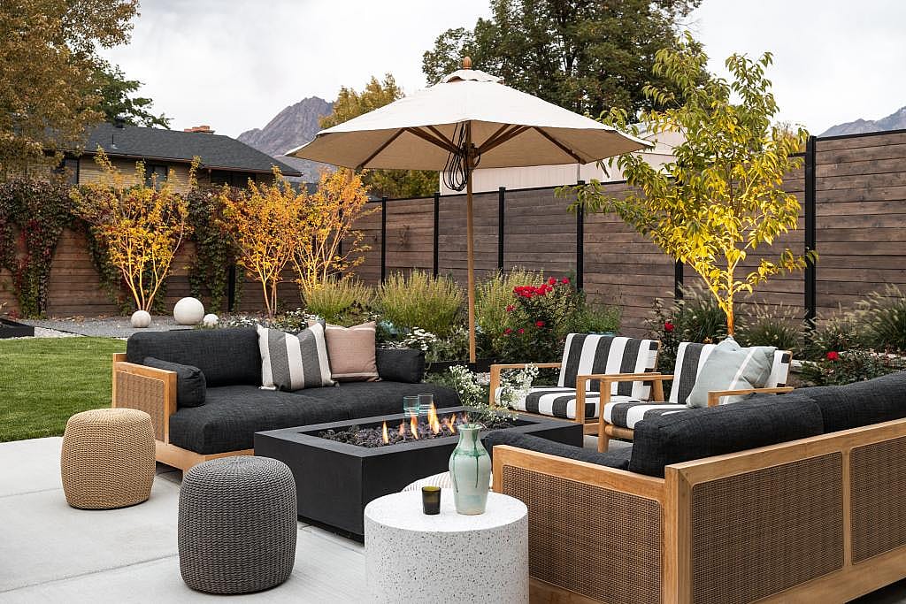 Backyard patio set in neutral black and tan colored furniture arranged around a low gas firepit and trees and horizontal wooden fence in the background.