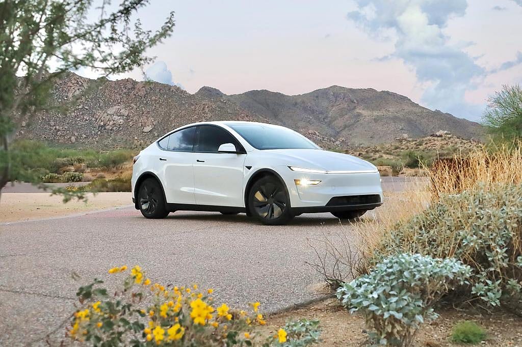 Front three-quarter view of white 2026 Tesla Model Y parked on pavement with native chaparral plants visible in foreground and background.