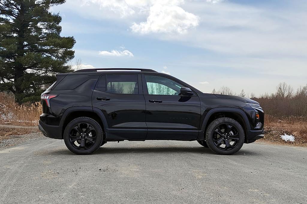 Side view of black 2026 Chevrolet Equinox parked on dirt road with sky and a pine tree in the background.