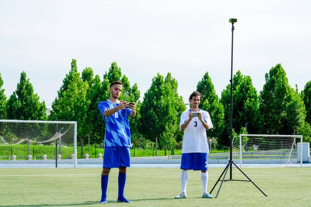 Two players using smartphones in a soccer field with the XbotGo Falcon 4K sports camera mounted on a tripod.