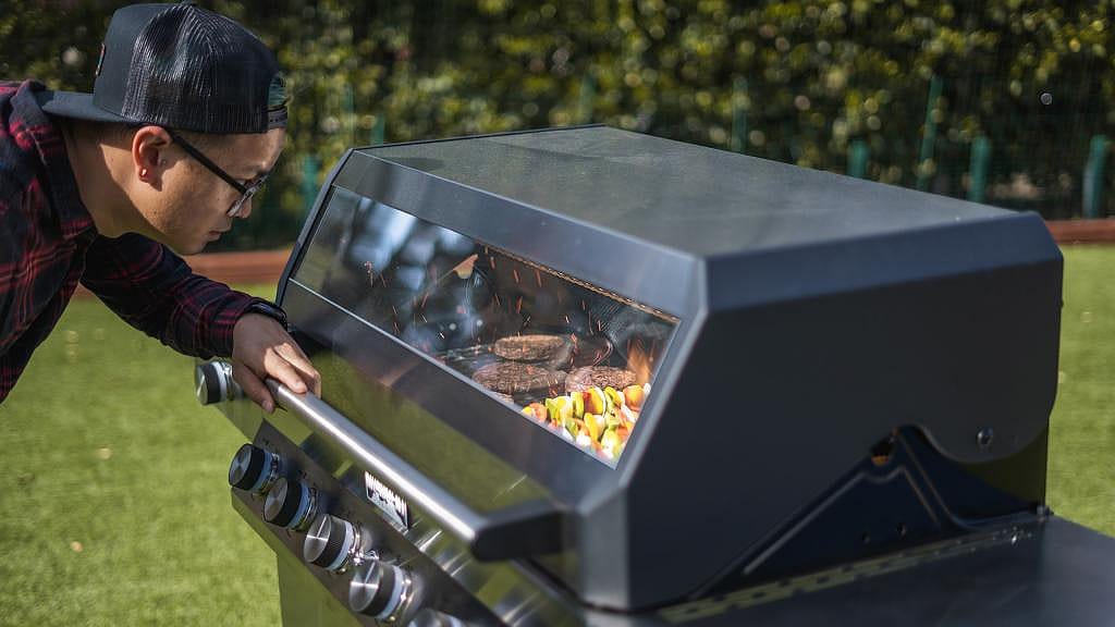 A person looking through the clear glass window of a modern black gas grill outdoors.