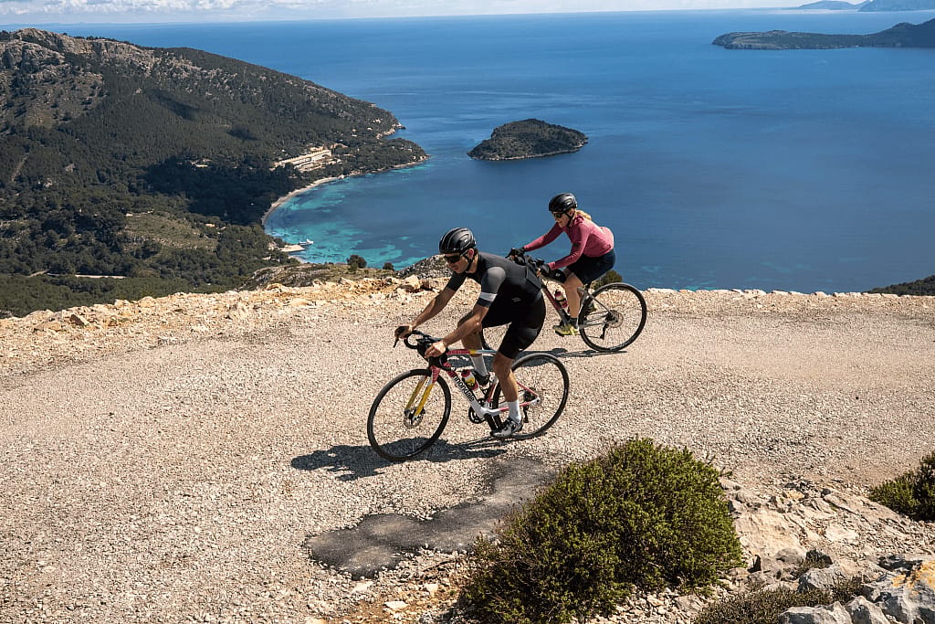 Two people biking on a wide gravel path near the ocean in Mallorca.