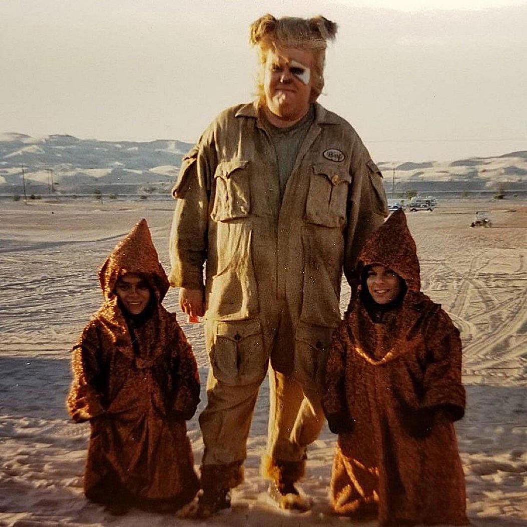 A large man in a khaki jumpsuit with a shaggy blonde wig and face paint stands in a desert landscape flanked by two small figures in hooded brown robes.