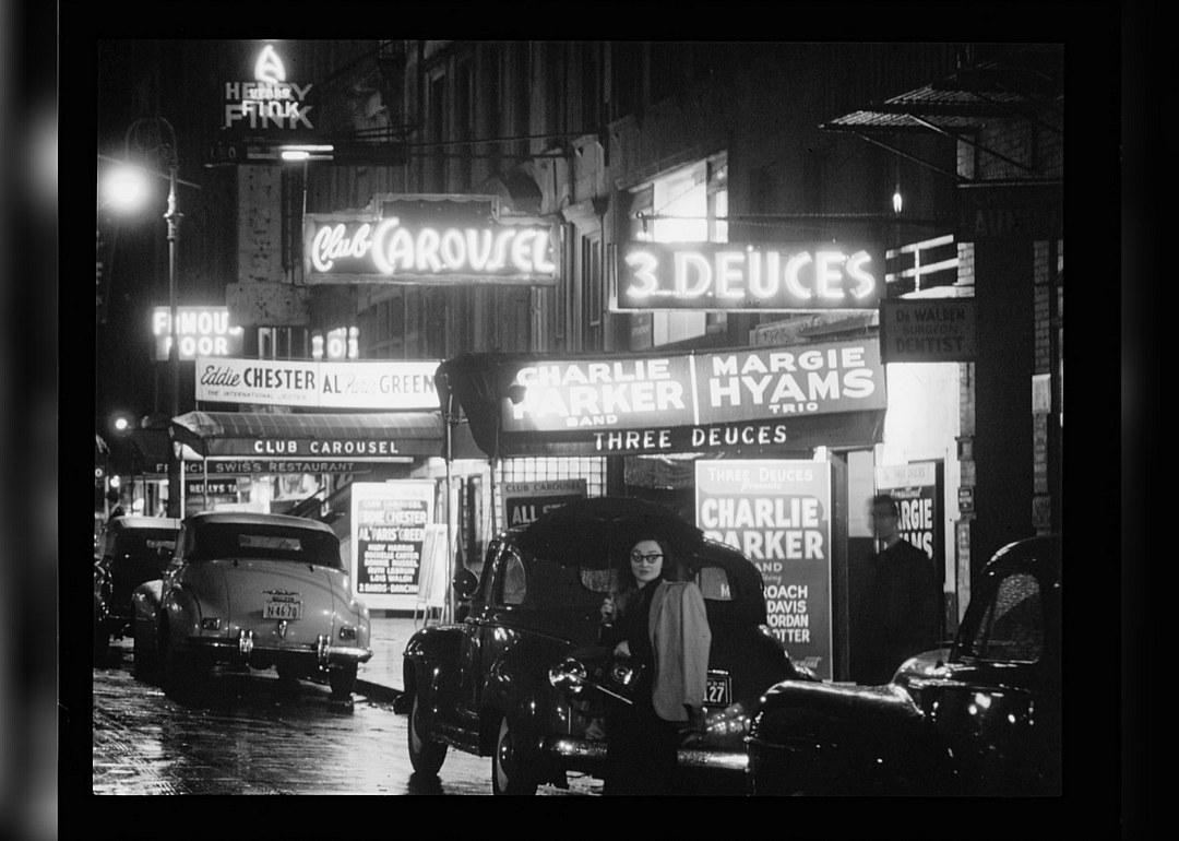 Neon-lit jazz clubs on 52nd Street in New York City at night, circa 1948, featuring Club Carousel and Three Deuces with vintage cars on the street