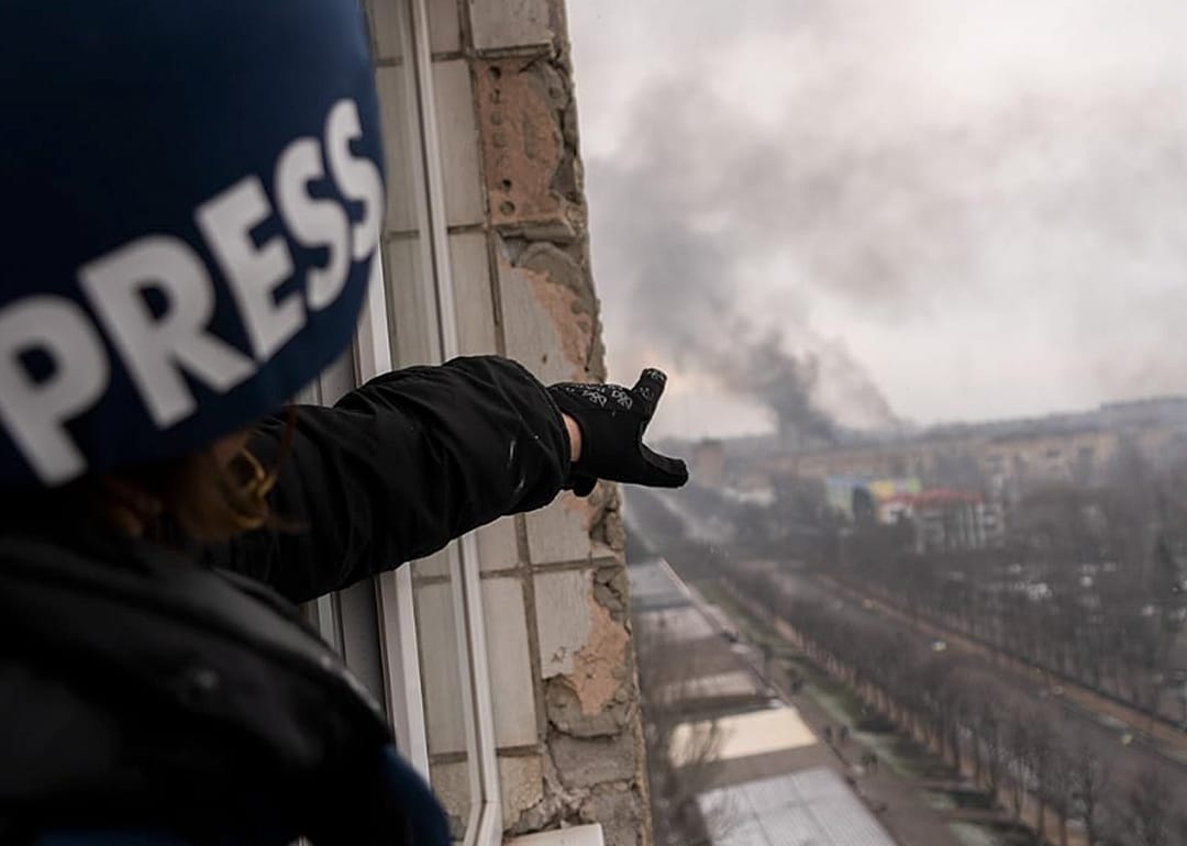 Person in a press helmet pointing to smoke rising over buildings.