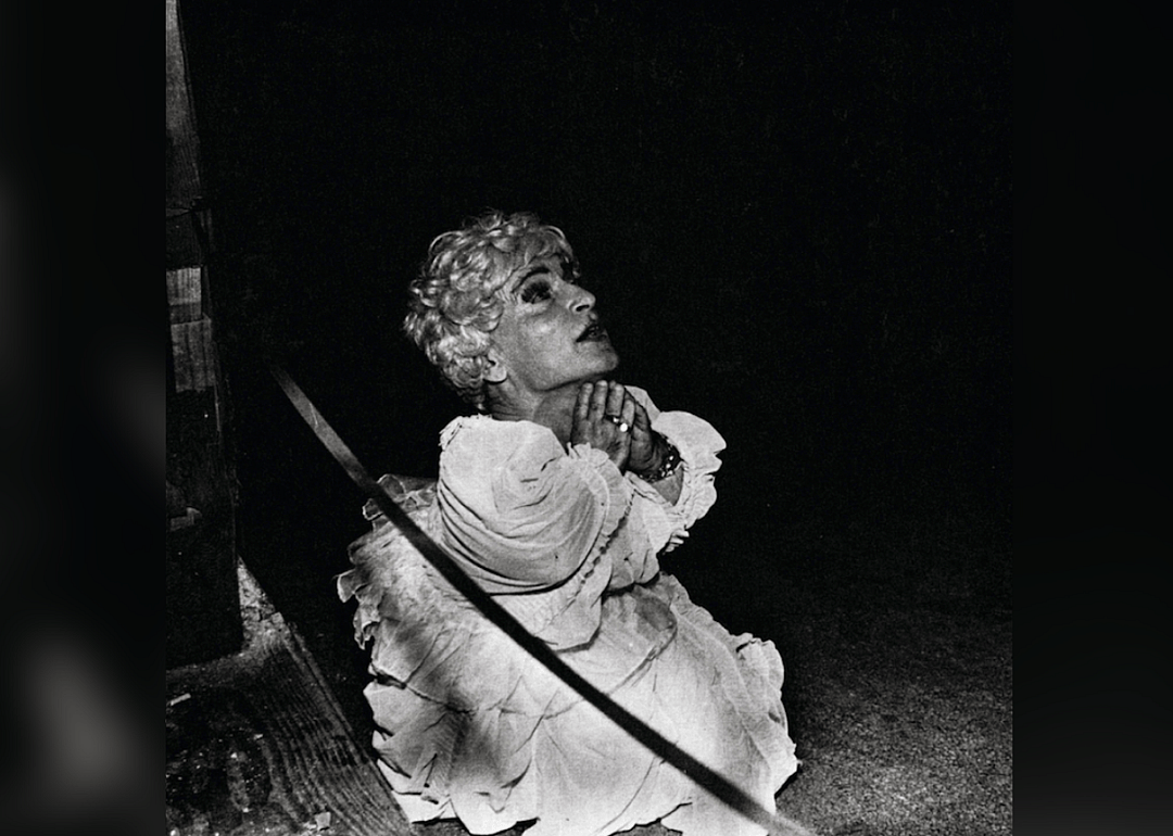 A little person in a dress and wig praying.