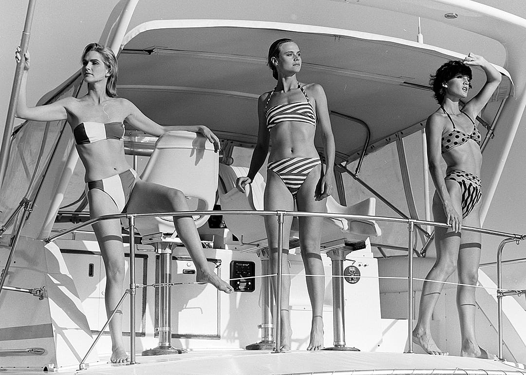 Three models pose on boat in swimwear.