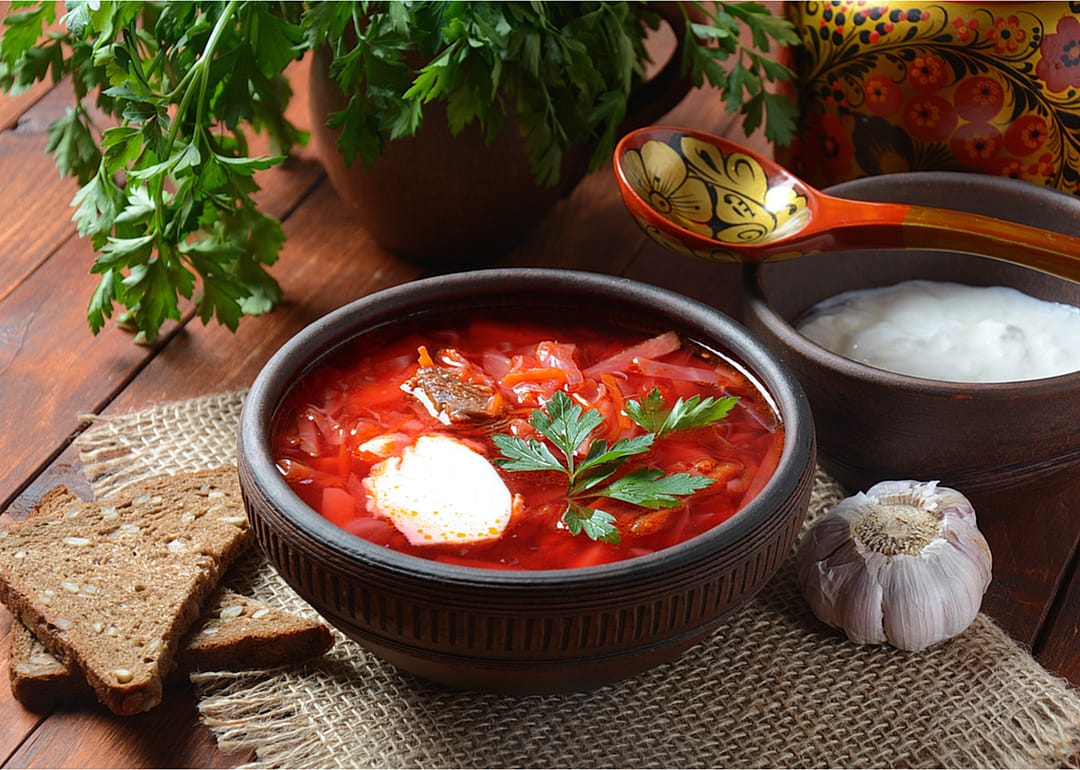 A bowl of borscht with slices of bread and a bowl of sour cream next to it, along with a bulb of garlic and a plant pot of parsley.