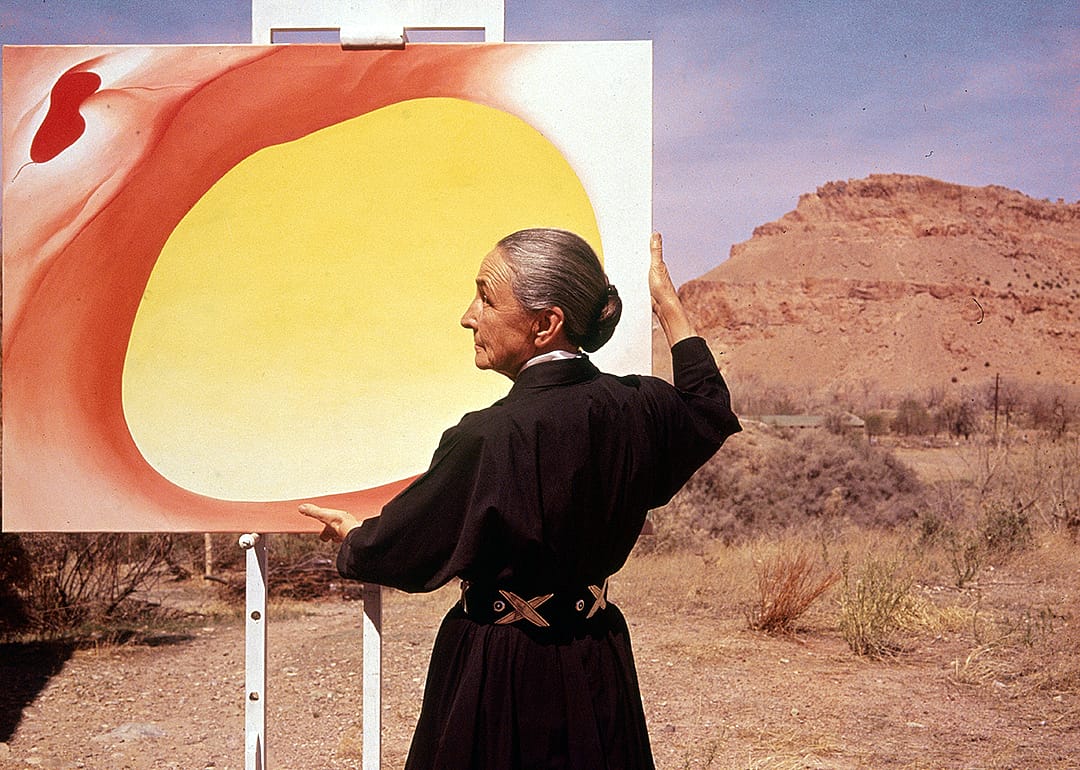 Georgia O'Keeffe stands at an easel with a large painting outdoors.