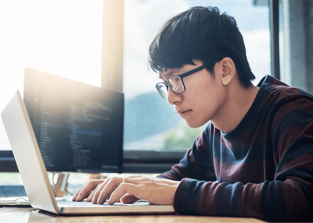 A young man with glasses looks skeptically at his laptop.