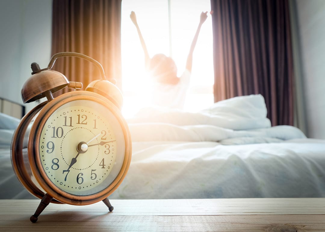 An alarm clock in the foreground and a waking teenager in the background.