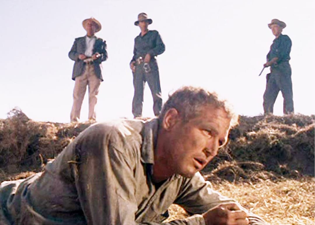 Paul Newman lies on the ground with three armed men watching him.