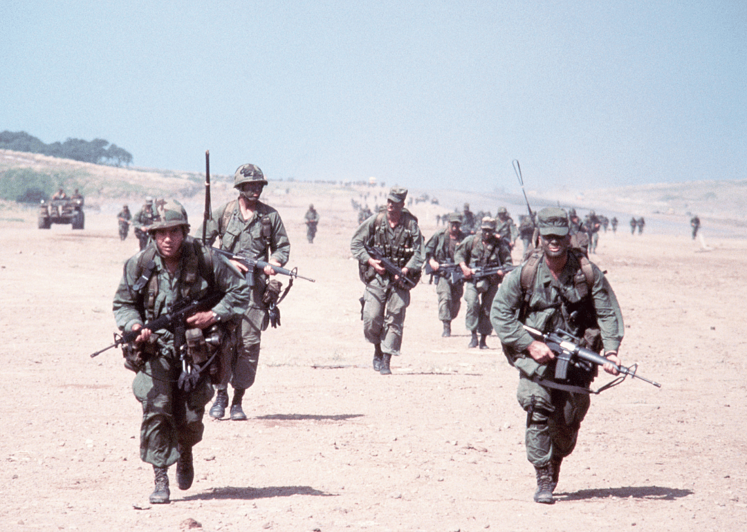 United States combat soldiers run in parallel lines across an empty dirt field during the invasion of the island of Grenada.