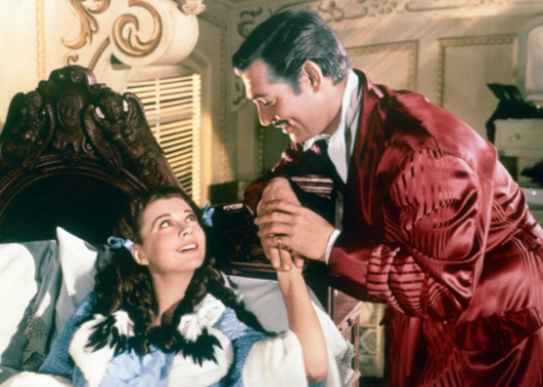 Clark Gable bending down to hold Vivien Leigh's hand as they gaze into each other's eyes.