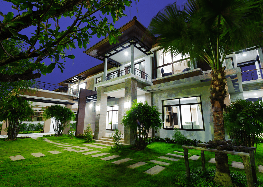 A white, modern mansion is illuminated from within in the evening.