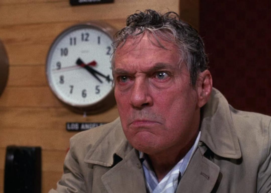 Peter Finch in a trench coat in front of a wall of clocks.