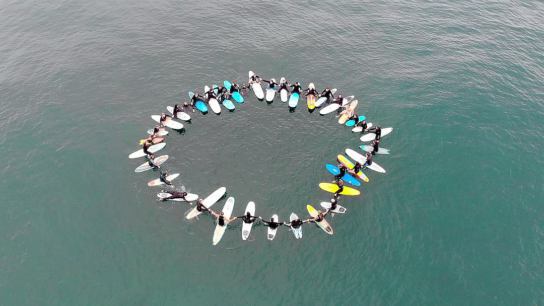 A group of surfers sit in a circle in the water on their boards