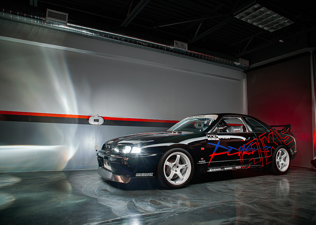 A modified black Nissan Skyline with its lights on in a parking area