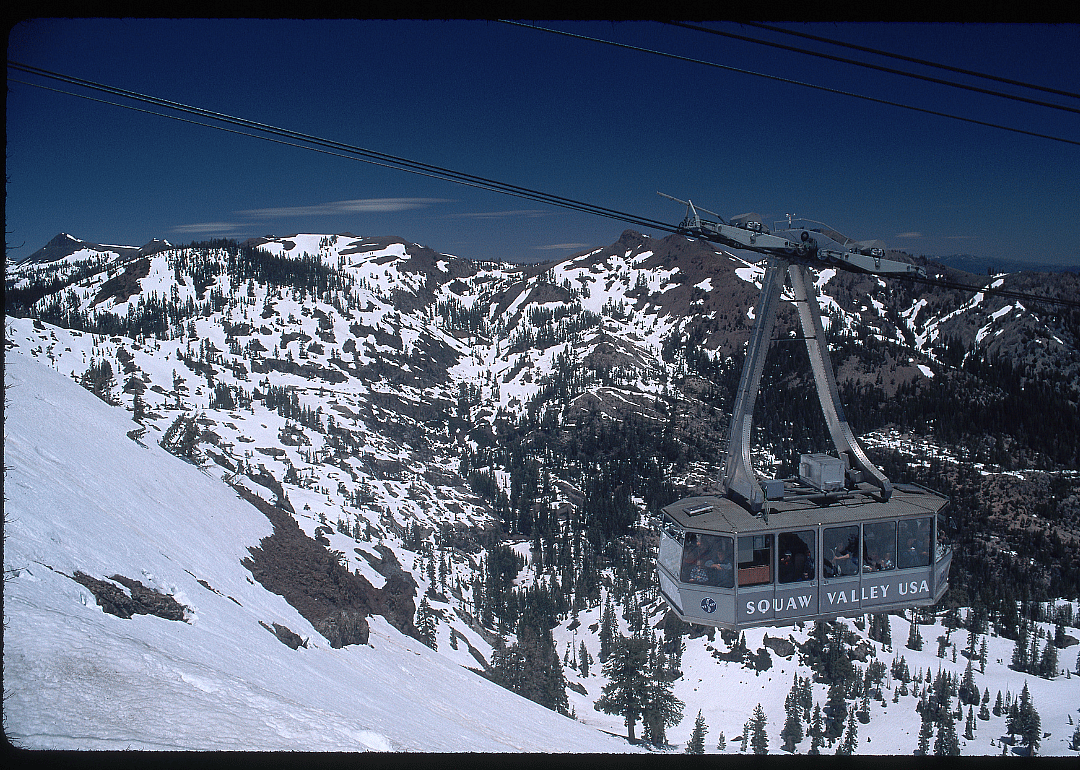 Exterior of an aerial tram at the Squaw Valley Ski Resort, with the Sierra Nevadas in the background. The tram is filled with skiers headed to the top of the mountain.