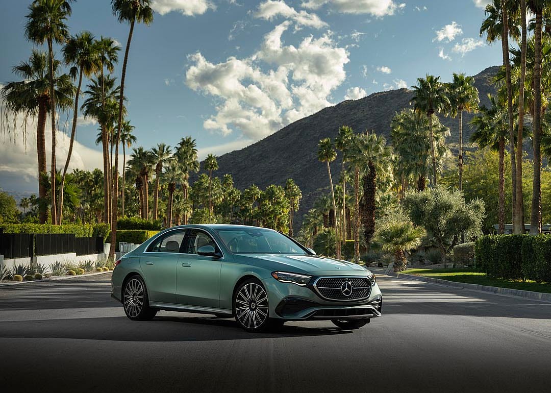 Teal Mercedes-Benz E-Class parked on road in front of desert oasis with palm trees in background.