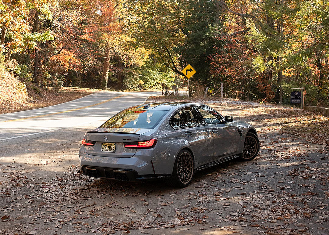 A metallic BMW M3 is parked on a leafy roadside.