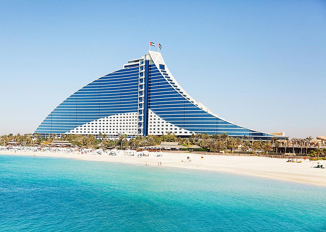 Jumeirah Beach hotel in Dubai with the pure white sand beach and crystal clear water in front of it surrounded by green palm trees.