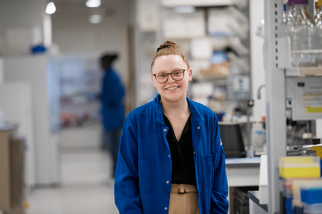 Madeline Szoo wearing a blue labcoat posing for a portrait in a lab.