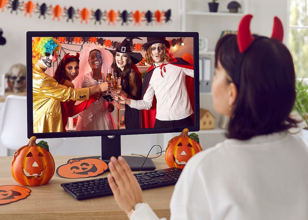 A woman, sitting in front of desktop computer at a table decorated for Halloween, greeting friends during a virtual Halloween costume party.