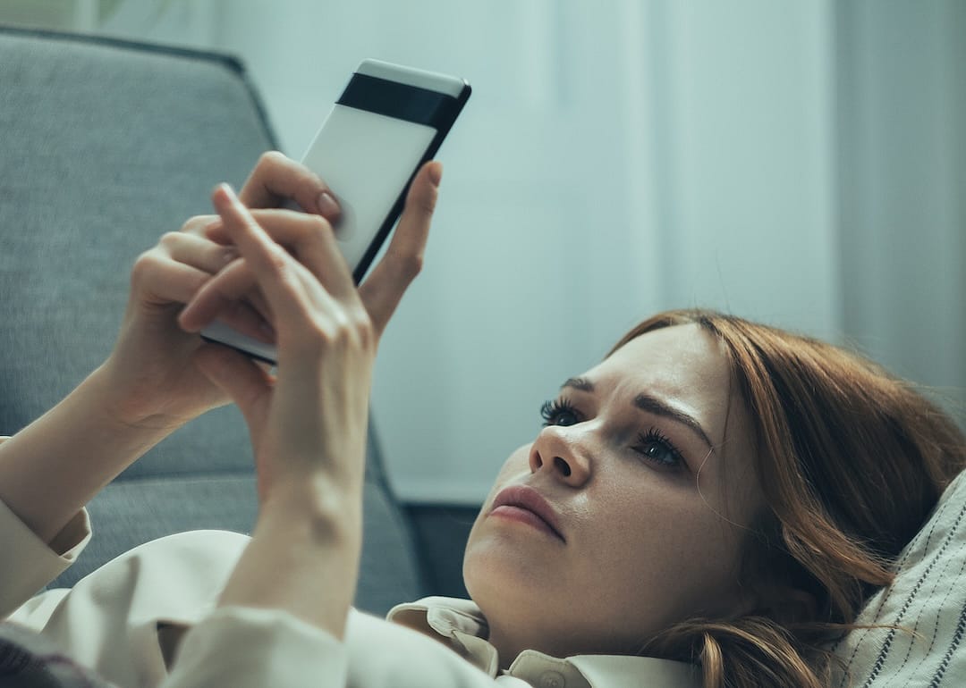 Girl lying with a phone in her hands and a concerned expression on her face as she looks at the screen.