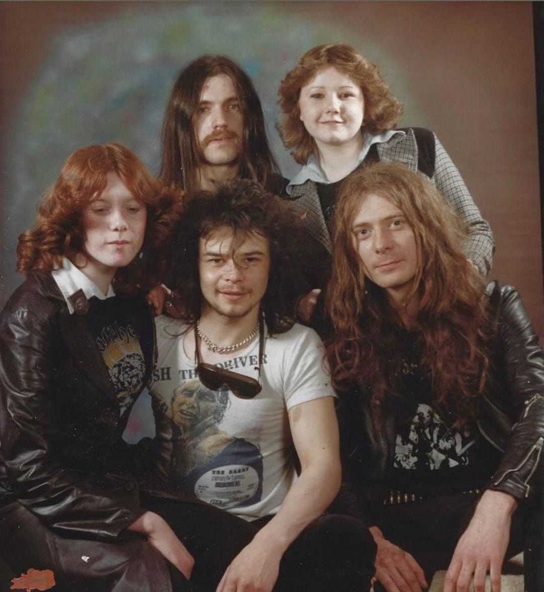 Color photo of five people posing together against a studio backdrop in 1977. Three are members of Motörhead, one tall man with long dark hair and a mustache standing at the back, alongside two young women with curly hair. Several wear band t-shirts and leather jackets.