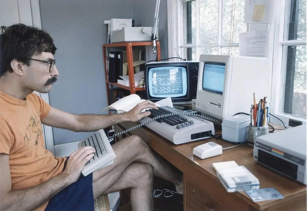 Man in an orange shirt sitting in front of two computers in the 1980s