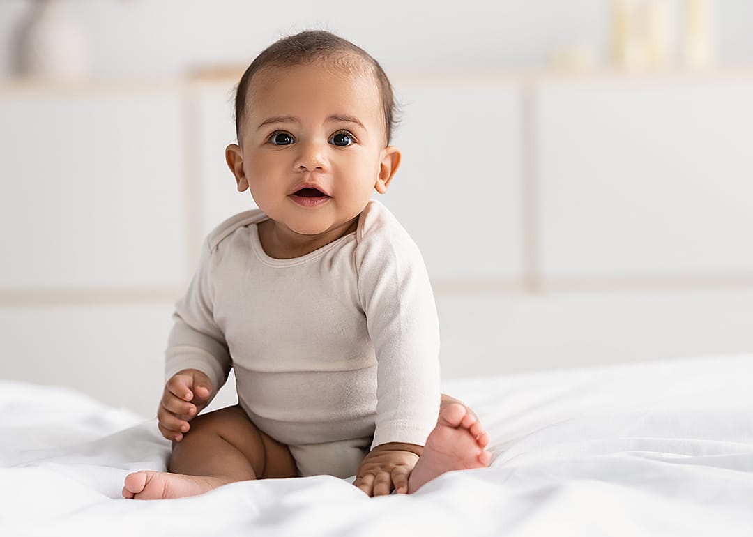 Baby wearing onesie sitting on bed.