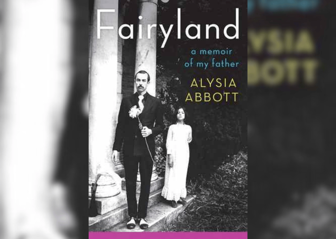 A black-and-white image of a little girl standing behind a man in a suit holding a flower.
