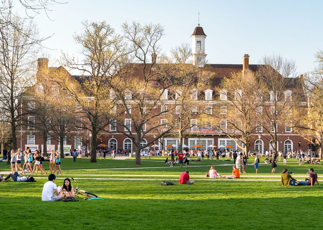 A large green lawn where people are lounging, sitting in groups. Some are sprawled on the grass with books while others are walking or cycling.In the background is a large, classic brick building with white-trimmed windows and a central cupola.