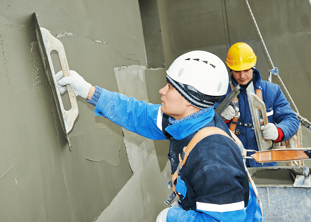 A man in a harness and white hard hat holds a tool while smoothing the concrete on the wall; another man in the background is seen holding tools. 