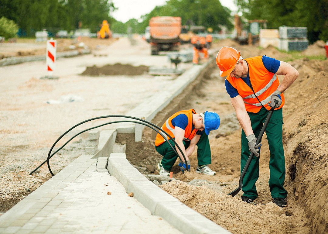 A man in an orange vest and hard hat holds a spade in the sand while another man in the background squats to hold black cables in the ground. 