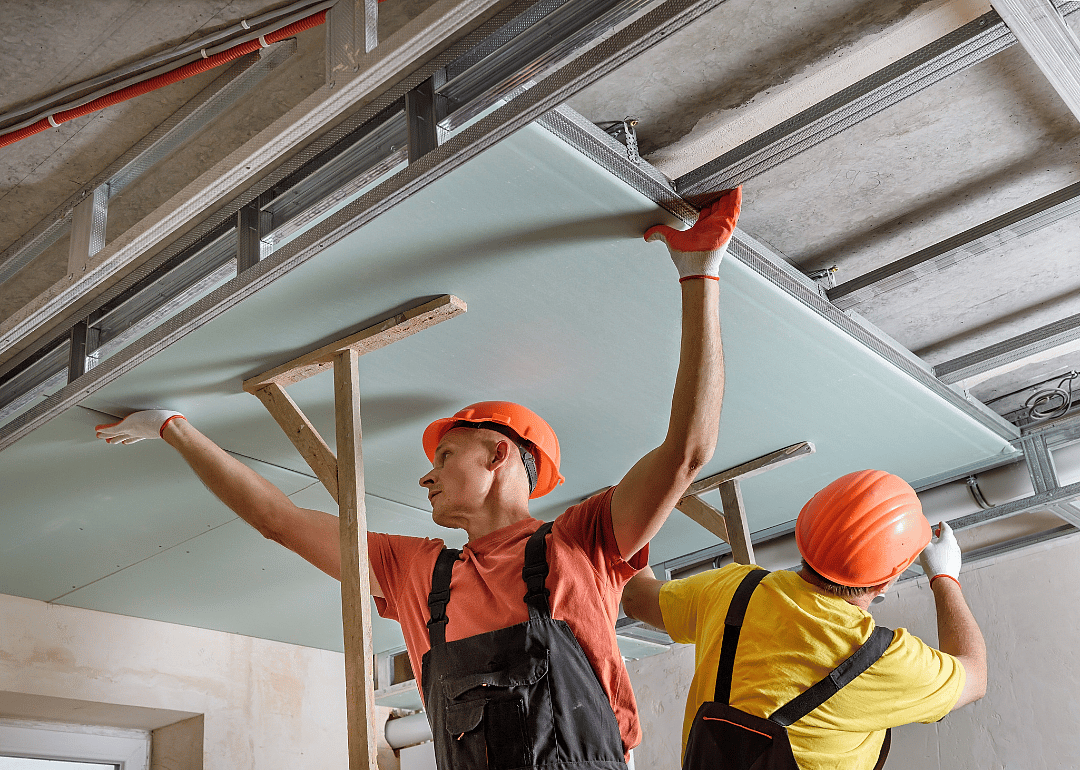 One man wearing a hard hat and overalls holds up a large piece of board in the ceiling, while another looks closer at the metal railing. 