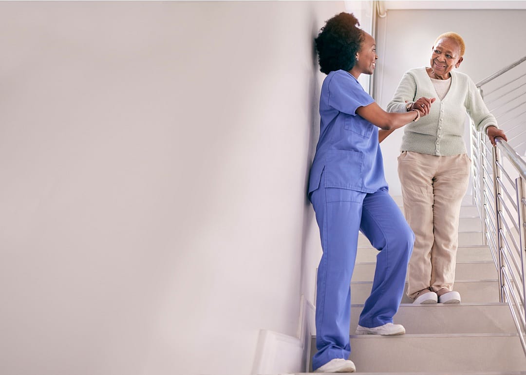 An elderly woman walking down the stairs with the help of a careworker in blue scrubs.
